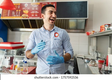 Young adult caucasian man entrepreneur standing at his fast food store in day smiling front view Chef in the kitchen having fun at work wearing protective medical gloves to prevent disease real people