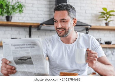 Young adult caucasian man reading news holding newspaper and drinking cup of tea coffee while having breakfast eating sandwich lunch brunch in modern kitchen in background - Powered by Shutterstock