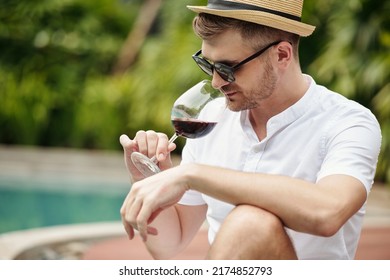 Young Adult Caucasian Man Wearing White Shirt, Hat And Sunglasses Sitting Relaxed At Poolside Taking Sniff Of Red Wine In Glass Before Tasting It