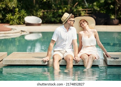 Young adult Caucasian man and woman in love enjoying spending time together at hotel territory sitting at pool with legs in water - Powered by Shutterstock