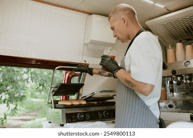 Young adult chef drinking cold brew coffee while checking buns for hot dogs on grill - Powered by Shutterstock