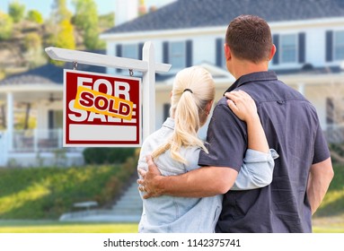 Young Adult Couple Facing Front Of Sold Real Estate Sign And House.