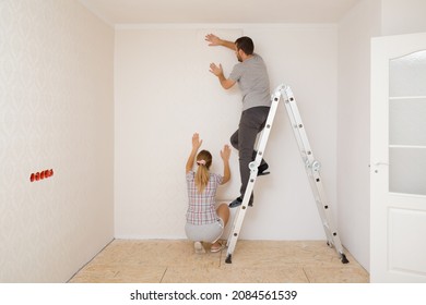 Young Adult Couple Making Interior Change. Man Standing On Metal Ladder And Woman Help Applying New Wallpaper On White Wall In Room. Working Together. Repair Work Of Home. 