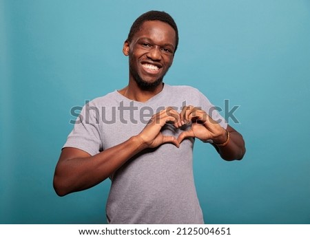 Young adult doing heart shaped sign with hands in studio, expressing amorous feelings on camera. Smiling man doing romantic gesture to express love, affection and passion for valentines day.