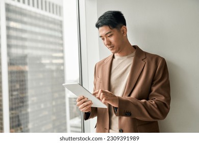 Young adult elegant professional Asian business man manager investor holding tablet using digital pc standing at office window doing digital data research on tech website analyzing financial market. - Powered by Shutterstock