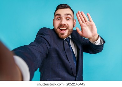 Young Adult Excited Satisfied Man Wearing Official Style Suit, Making Selfie Or Streaming, Waving Hand POV, Point Of View Of Photo. Indoor Studio Shot Isolated On Blue Background.