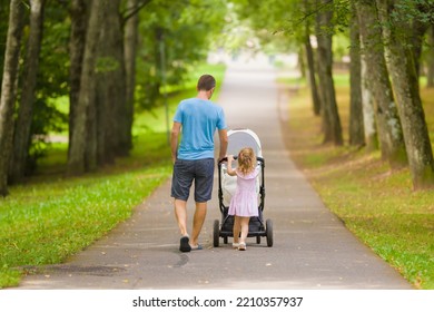 Young Adult Father And Little Daughter Pushing White Baby Stroller And Walking At Town Park In Summer Day. Spending Time Together And Breathing Fresh Air. Enjoying Stroll. Two Child Dad. Back View.