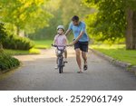 Young adult father teaching happy beautiful little girl to ride on first bike on sidewalk at city park. Learning to keep balance. Warm summer day. Cute 5 years old toddler. Front view.