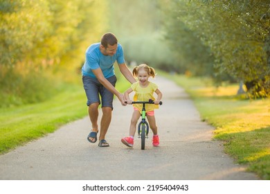 Young Adult Father Teaching Happy Beautiful Little Girl To Ride On First Bike Without Pedals On Sidewalk At City Park. Learning To Keep Balance. Warm Summer Day. Cute 3 Years Old Toddler. Front View.