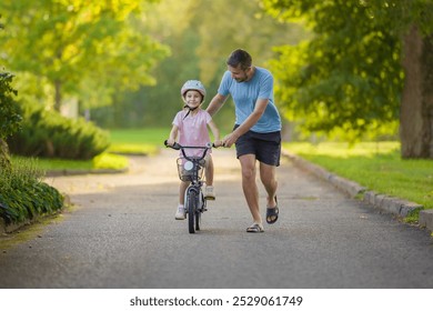 Young adult father teaching happy beautiful little girl to ride on first bike on sidewalk at city park. Learning to keep balance. Warm summer day. Cute 5 years old toddler. Front view. - Powered by Shutterstock