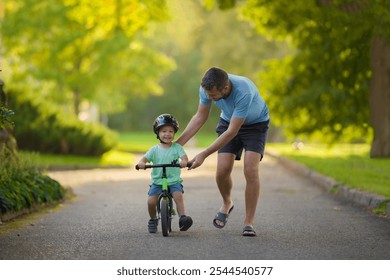 Young adult father teaching happy beautiful little boy to ride on first bike without pedals on sidewalk at city park. Learning to keep balance. Warm summer day. Cute 2 years old toddler. Front view. - Powered by Shutterstock