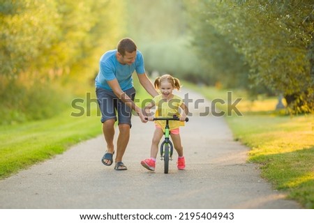 Young adult father teaching happy beautiful little girl to ride on first bike without pedals on sidewalk at city park. Learning to keep balance. Warm summer day. Cute 3 years old toddler. Front view.