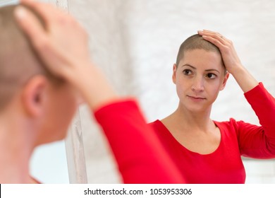 Young Adult Female Cancer Patient Looking In The Mirror, Stroking Her New Short Hair. 