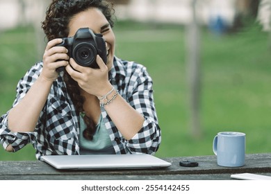 Young adult female photographer smiling while capturing images with a dslr camera in a park, laptop and coffee on the table, embracing her passion for photography - Powered by Shutterstock