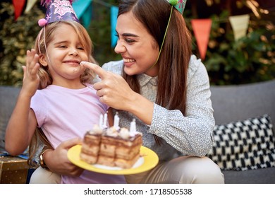 Young Adult Female Putting Whipped Cream From A Birthday Cake On A Nose Of A Cute Little Girl