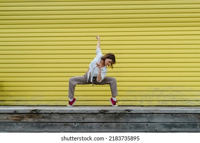 Young Adult Female In White Shirt And Grey Linen Pants Dancing In Front Of Bright Yellow Wall, Selective Focus