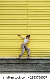 Young Adult Female In White Top And Grey Pants Dancing In Front Of Bright Yellow Wall. Selective Focus