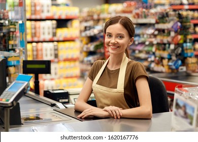 Young Adult And Friendly Woman Cashier With Smiling And Cheerful Face Sitting Behind Checkout In Market, Looking At Camera