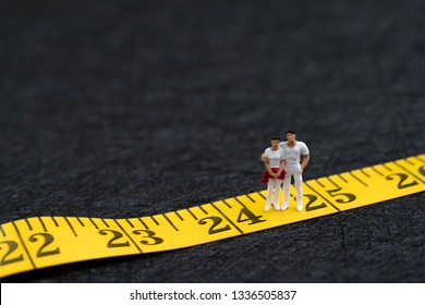 Young Adult, Gen Y Or New Age People Concept, Miniature People Couple In Casual White Cloth Standing On Number 25 On Yellow Measuring Tape Metaphor Of Their Age.