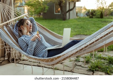 Young Adult Girl In A Hammock Works On A Computer Outside The House And Smokes An Electronic Cigarette. 