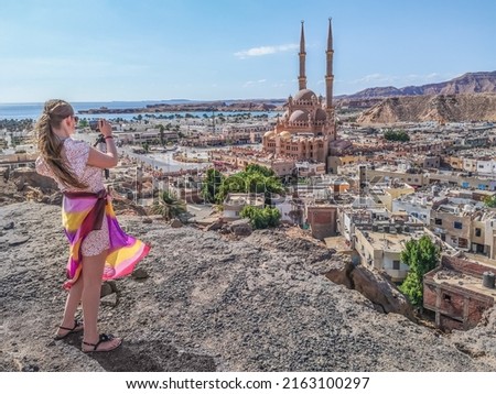 Young adult girl takes pictures of Al Sahaba Mosque in the middle of Old Market in Sharm El Sheikh, standing on top of a cliff. European woman on the backdrop of an exotic panorama of Egyptian city