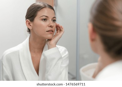 Young adult and good-looking woman in white bathrobe standing in bright light bathroom with mirror. She looking at her reflection with calm face - Powered by Shutterstock