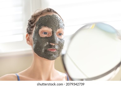 Young Adult Happy Beautiful Caucasian Woman Applying Natural Gray Clay Face Mask At Home Against Window And Mirror At Day Time. Female Person Making Self Facial Cleansing Moisturizing Treatment Care