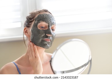 Young Adult Happy Beautiful Caucasian Woman Applying Natural Gray Clay Face Mask At Home Against Window And Mirror At Day Time. Female Person Making Self Facial Cleansing Moisturizing Treatment Care