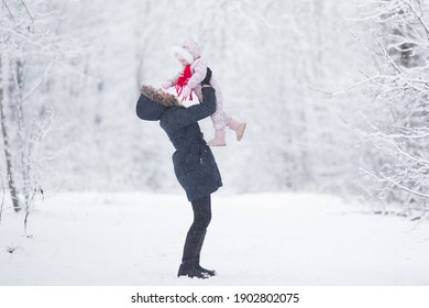 Young Adult Happy Mother Lifting Up Smiling Baby Girl At White Snowy Park After Blizzard. Looking At Each Other. Side View. Spending Time Together In Beautiful Winter Day. Lovely Emotional Moment.