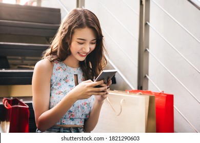 Young Adult Happy Smile Asian Woman Using Mobile Phone For Online Application. Sitting With Shopping Bags Background. City Lifestyle People Concept.