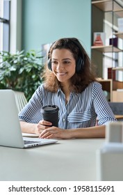 Young Adult Happy Smiling Hispanic Indian Student Wearing Headphones Talking Online Chat Meeting Holding Coffee Using Laptop In Virtual Office. College Female Student Learning Remotely.