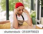 Young adult hispanic barista is sitting at a table in a coffee shop, wearing an apron and a red beanie, massaging his temples with a painful expression