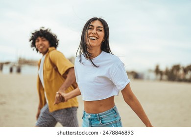 A young adult Hispanic couple in casual clothes revel in their beach day, laughing together with windswept hair as they playfully run on the sand. - Powered by Shutterstock