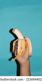 young adult hispanic hand holding a peeled and bitten banana on blue background 