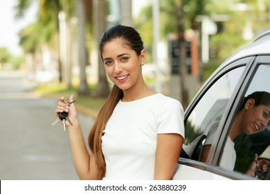 Young Adult Hispanic Woman Holding Keys Of New White Car, Leaning On Auto Door. She Smiles And Looks Proud At Camera, Showing The Keys. Waist Up Portrait