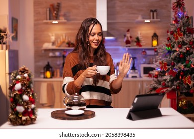 Young Adult Holding Cup Of Tea While Waving On Online Chatting Video Call Conference With Friends. Caucasian Woman Using Wireless Internet Technology On Tablet In Christmas Decorated Home