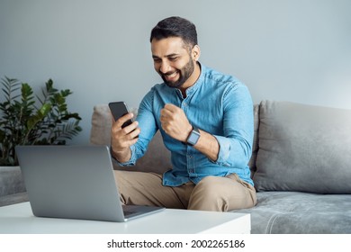 Young Adult Indian Business Man Screaming Proud Cheering With Excitement Reading Great News On Smartphone Celebrating Victory And Success Sitting Front Of Laptop Computer Working At Home Office