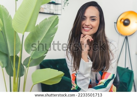 Young adult latin american woman wearing shirt with fun colorful pattern sitting in modern living room next to plant posing for camera with friendly smile. Horizontal indoor shot. High quality photo