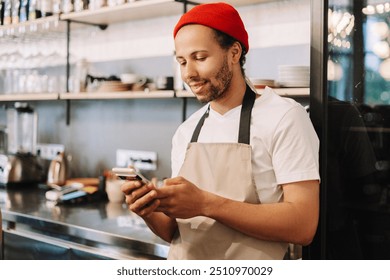 Young adult male barista wearing a red beanie and an apron is checking his phone and smiling while working behind the bar at a cafe - Powered by Shutterstock