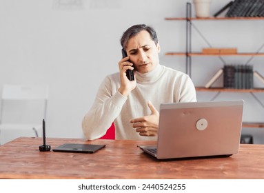 Young adult male graphic designer talking on a mobile phone in the office and working on a laptop and graphics tablet. - Powered by Shutterstock