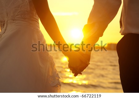 Young adult male groom and female bride holding hands on beach at sunset.