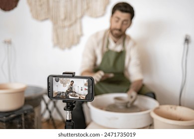 Young adult male potter sculpting vase on potter's wheel while recording video blog using smartphone on tripod. - Powered by Shutterstock