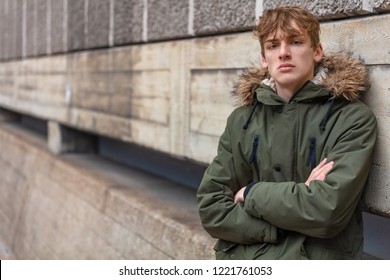 Young Adult Male Teenager Boy Outside In An Urban City Wearing A Green Parka Coat Leaning Against A Wall