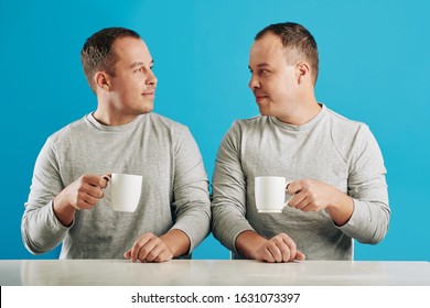 Young adult male twin siblings sitting at table looking at each other holding cups with coffee, blue background
