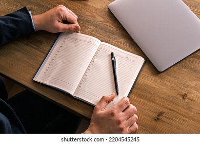 Young Adult Male Writing Down Notes On A Notebook. Unrecognizable Businessman With Laptop Computer On The Desk Working. Education, Study Or Work.