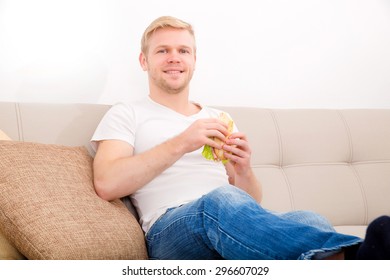 A Young Adult Man Eating A Sandwich At Home On The Couch.
