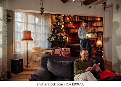 Young Adult Man With Eyeglasses Relaxing On Sofa And Using Digital Tablet While His Wife Approaches From Behind With Christmas Gift