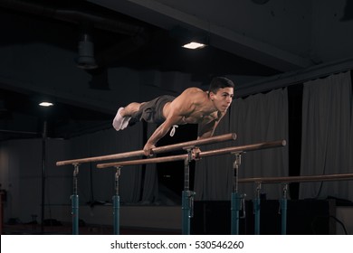 Young Adult Man Gymnast In Air, Holding Gymnastic Rings. Indoors, Dark Hall, Gymnastics Equipment.