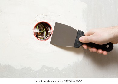 Young Adult Man Hand Using Spatula And Plastering Wall With White Fresh Putty Around New Plastic Outlet. Closeup. Repair Work Of Home.