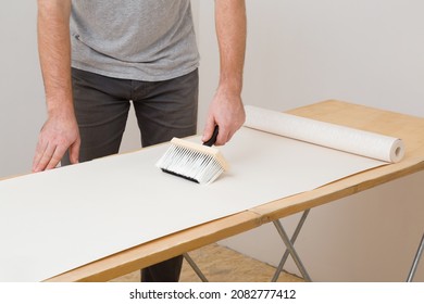 Young Adult Man Hand Using Brush And Applying Glue On Wallpaper Sheet On Wooden Table. Closeup. Repair Work Of Home. 